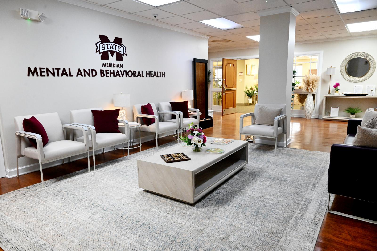 Photo showing waiting room in the new Mental and Behavioral Health Clinic at MSU-Meridian. White carpet with white chairs and maroon pillows with white coffee table. Maroon MState logo on the wall with maroon letters beneath "MERIDIAN MENTAL AND BEHAVIORAL HEALTH"