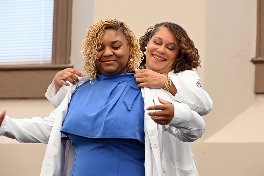 Mississippi State University - Meridian, Master of Physician Assistant Studies Program White Coat Ceremony. Student wearing blue dress is being ceremonially helped into a white medical lab coat by professor also wearing a white medical lab coat.